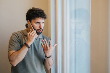 A man standing near a window making a phone call, exhibiting concern or questioning gestures,...
