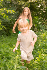 A happy blonde boy with long hair is running along the path with his blonde sister in a green sundress
