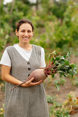 A cheerful, smiling woman proudly displays her freshly harvested beetroot amidst her vibrant and lush garden setting