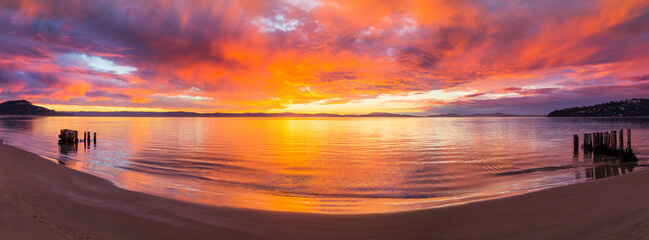 Panorama view of a colourful sunrise reflected in a calm ocean bay with wooden groynes