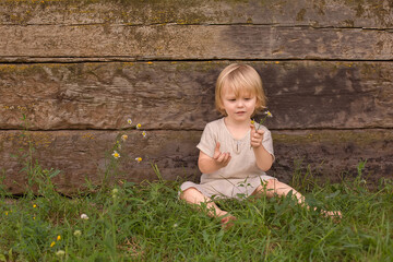 A happy blond boy with long hair is sitting on the grass by a wooden wall with a flower in his hands