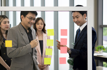 A professional man and woman stand together, engaged in a brainstorming session, using document and colorful paper note on a transparent glass wall.