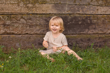 A happy blond boy with long hair is sitting on the grass by a wooden wall with a flower in his hands