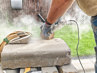 Worker uses tool to cut stone block in outdoor workspace during sunny day