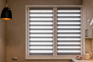 Day-night dual roller blinds on kitchen window, made of double fabric layers with alternating stripes. It's a teapot and a vase with peaches on  countertop. Zebra double roller shades in the interior.