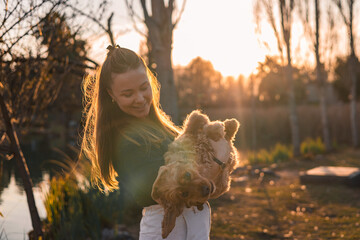 A young Caucasian woman with long brown hair holds a small golden cocker spaniel in a sunlit...