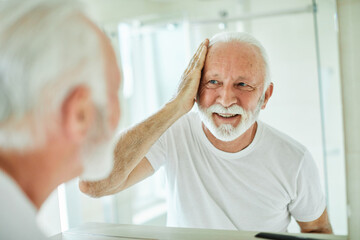 Portrait of an elderly senior man grooming and combing his beard and hair in front of  mirror in...