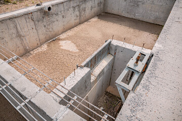 Empty concrete water tank during construction phase, featuring cracked soil, metal sluice gate, and reinforcement bars