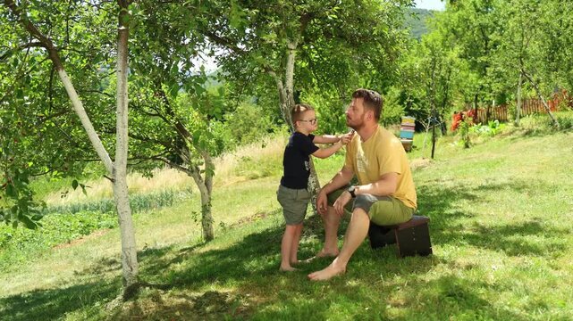 Cute boy with Down syndrome trimming his father's beard in the yard. Summer, nature, country life, people with disabilities inclusion. Active disabled persons, candid family. Real, authentic people. 
