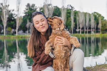 A young Caucasian woman with long brown hair smiles while holding a golden cocker spaniel by a...