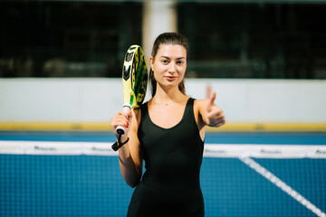 Young woman smiles and gives thumbs up after padel match at indoor court in the evening
