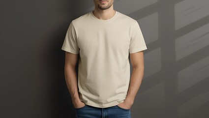 Man in a simple beige t-shirt standing against a dark gray wall with his hands in his pockets, looking  straight ahead in an indoor setting  wall, dark, indoor, person, casual, clothing, apparel, fash