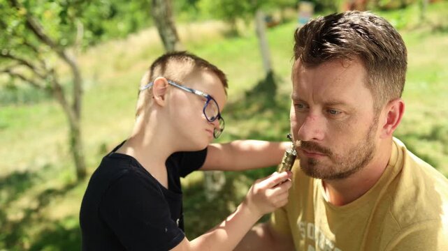Close up shot of a cute boy with Down syndrome and his father in the backyard. Boy is trimming his father's beard.summer, family, vacation, nature, real authentic people. Candid diverse people. Family