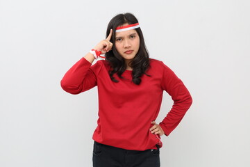 Indonesian woman with red and white ribbon celebrating Indonesian independence day, serious expression, thinking gesture while holding forehead, standing looking at camera.