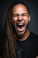 A man with long dreadlocks passionately screams with eyes closed against a dark background.