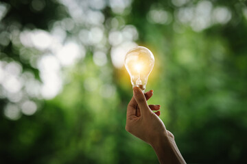 A hand holds a glowing light bulb against a green, blurred background, symbolizing innovation,...