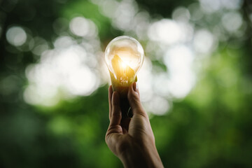 A hand holds a glowing light bulb against a green, blurred background, symbolizing innovation,...