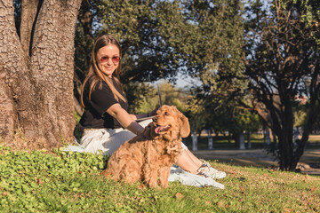 A young Caucasian woman with long brown hair sits on the grass beside a golden cocker spaniel dog...