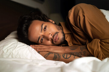 A young hispanic man with visible tattoos lies comfortably on a bed, gazing thoughtfully into the distance in a softly lit room. Generative AI.