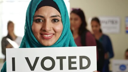 A smiling woman wearing a head covering holds a sign indicating she participated in a democratic voting process, encouraging engagement.