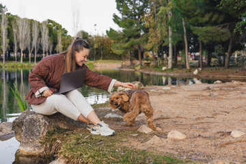 A young Caucasian woman with long brown hair sits by a lake, playing with a small brown cocker...