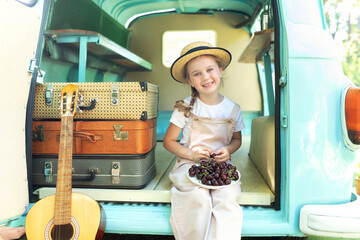 Girl in a straw of a hat and overalls sit near on the doorstep of the motor home and eating cherries. Smile child girl near old hippie bus in Campsite with guitar, suitcases holding fresh fruits. 
