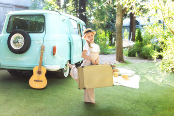 Girl in a straw of a hat and overalls with on suitcase in hands near of the motor home. Smile child girl near old hippie bus in Campsite with guitar. Concept family vacation, trip by car, campervan rv