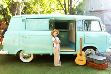 Girl in a straw of a hat and overalls standing near on the doorstep of the motor home. Smile child girl near old hippie bus in Campsite with guitar, suitcases. Concept vacation, trip by car, campervan