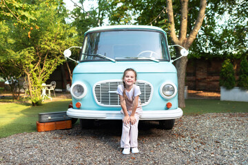 Emotion portrait cute fun Girl in overalls standing near of the motor home. Smiling child girl near old blue hippie bus in Campsite. Concept family vacation, trip by car, campervan. Kid in camping
