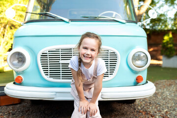 Emotion portrait cute fun Girl in overalls standing near of the motor home. Smiling child girl near old blue hippie bus in Campsite. Concept family vacation, trip by car, campervan. Kid in camping