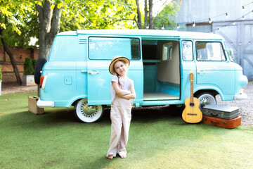 Girl in a straw of a hat and overalls standing near on the doorstep of the motor home. Smile child girl near old hippie bus in Campsite with guitar, suitcases. Concept vacation, trip by car, campervan