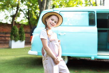 Cute fun Girl in a straw of a hat and in overalls standing near of the motor home. Smiling child girl near old hippie bus in Campsite. Concept family vacation, trip by car, campervan. Kid in camping