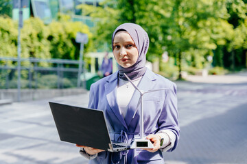 Muslim woman in lilac outfit holding laptop and wind turbine model outdoors