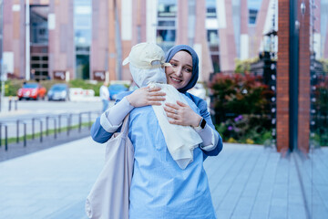 Muslim women hugging joyfully on city sidewalk in front of modern building