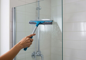 Hand cleaning a glass shower door with a squeegee in a modern bathroom setting during daylight