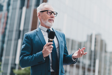 Mature professional man presenting outdoors in a formal suit holding a microphone, expressing...