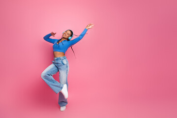 Smiling young woman wearing stylish blue top and jeans dancing happily with headphones against a...