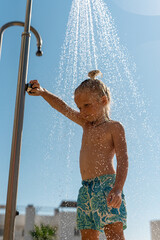 Young blonde boy with long hair taking a refreshing shower outdoors on a sunny summer day, wearing...