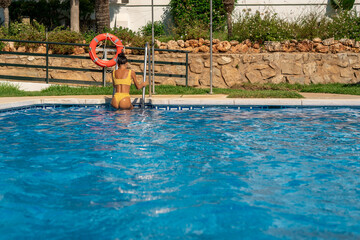 Athletic woman in a yellow bikini climbing out of a clear blue swimming pool using a metal ladder,...