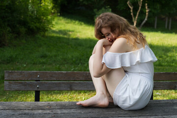 A young girl in a white off-shoulder dress sits thoughtfully on a wooden bench in a tranquil grassy park, surrounded by trees, immersed in quiet contemplation and natural serenity
