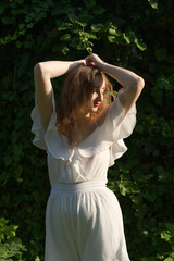 A woman in a white dress with ruffled sleeves poses gracefully against a backdrop of lush green foliage, bathed in warm sunlight.
