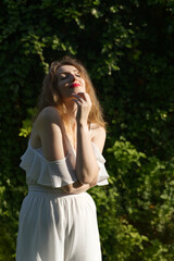 A young woman stands gracefully outdoors in an off-shoulder white top and pants, surrounded by lush greenery, with dappled sunlight casting soft shadows across her serene, stylish presence.