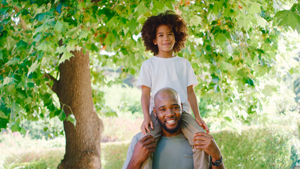 Portrait Of Father And Son In Summer Garden With Boy Riding On Dads Shoulders