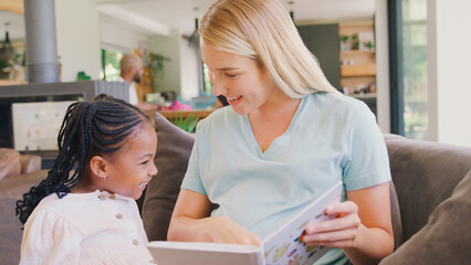 Mother And Daughter Reading Book At Home Together With Family In Background