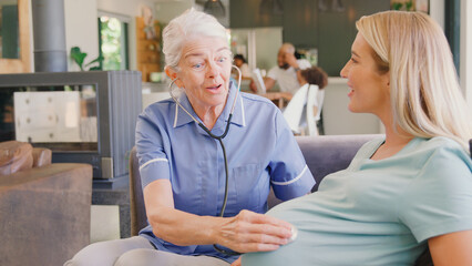 Senior Midwife With Stethoscope Visiting Pregnant Woman At Home With Family In Background