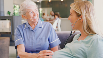 Senior Midwife With Stethoscope Visiting Pregnant Woman At Home With Family In Background