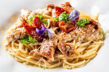Close-up of gourmet pasta with grilled chicken, sun-dried tomatoes, Parmesan, fresh herbs, and edible flowers on a white plate.