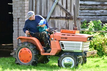 Grandfather and grandson on small tractor outside barn. Elderly man teaches young boy to steer. A...