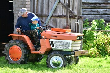 Grandfather and grandson on small tractor outside barn. Elderly man teaches young boy to steer. A...