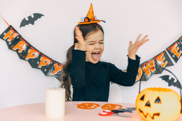 Happy emotional little girl dressed in a witch costume, prepares for Halloween by making...